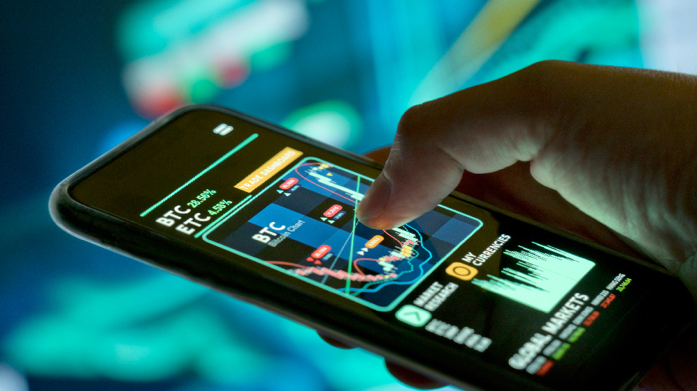 تصویر یک گوشی موبایل در دست یک نفر که نمودارهای ارز دیجیتال را نشان می‌دهد